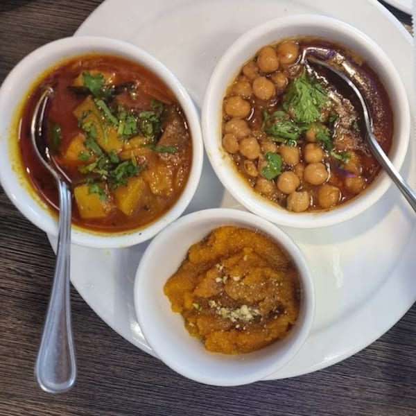 Three small white bowls of Pakistani food: two curries with chickpeas and vegetables, and one sweet halwa.